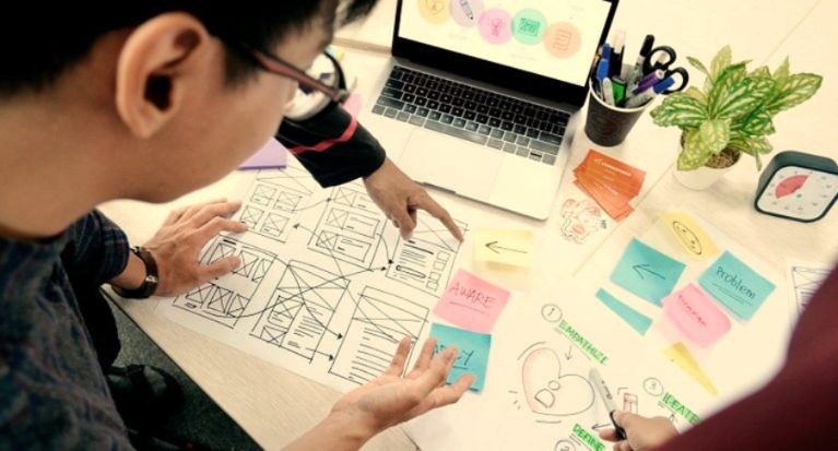 Bid writer examining a complicated bid process flow while another points at one element, surrounded by notes and a laptop.